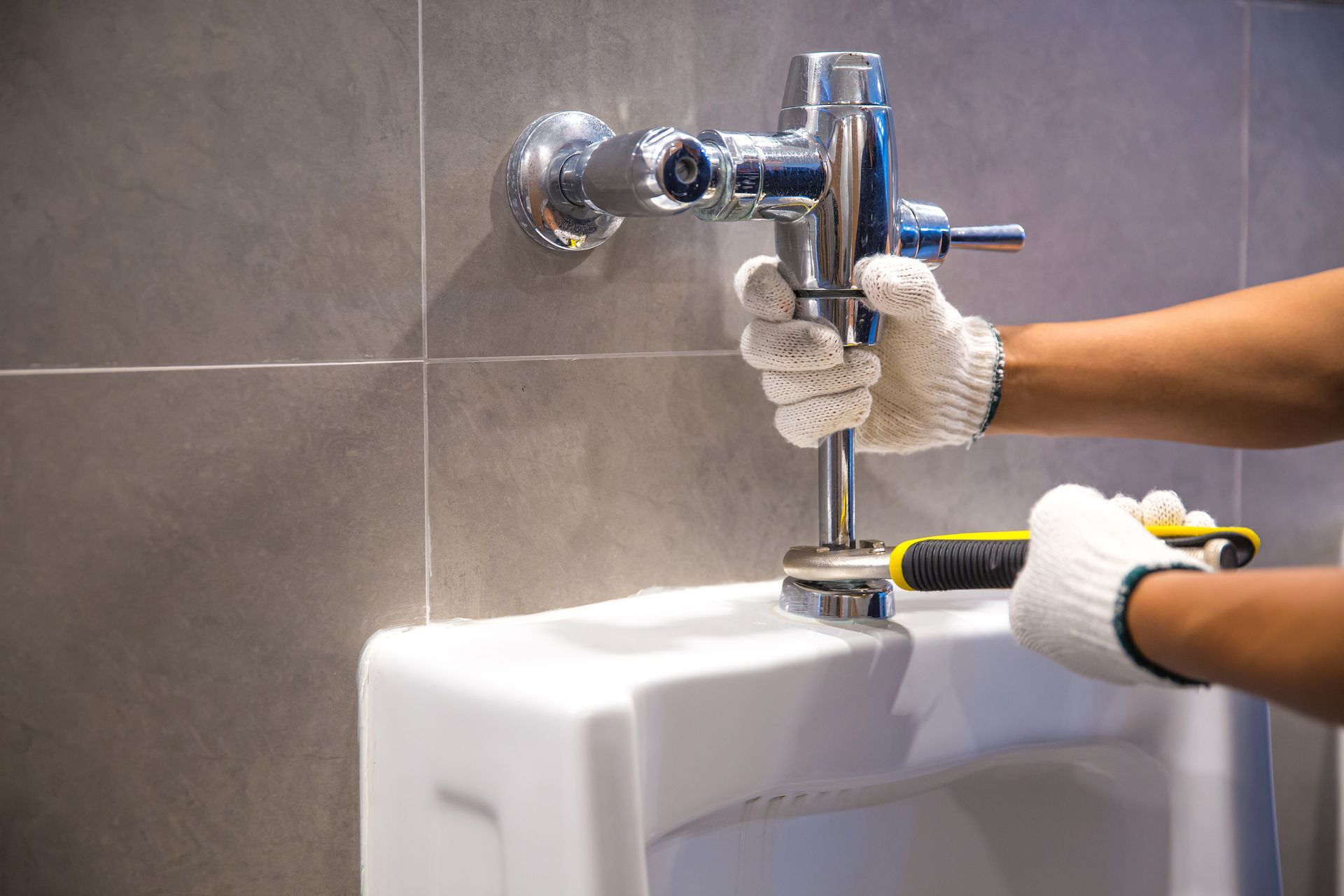 Gloved hands tightening a flush valve on a white urinal with a wrench in a tiled restroom