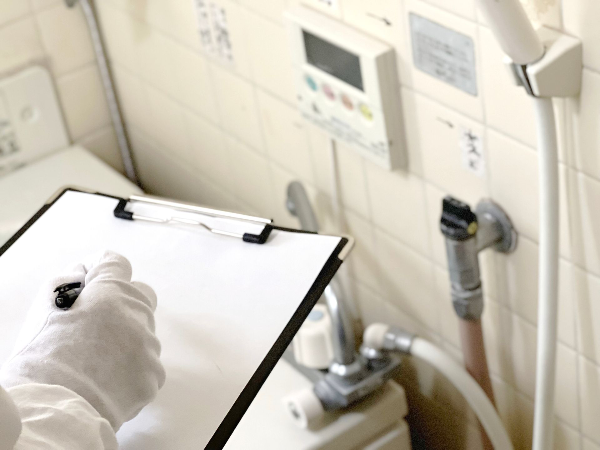 Gloved hand holding clipboard in a tiled bathroom or clinical washroom near a sink and pipes