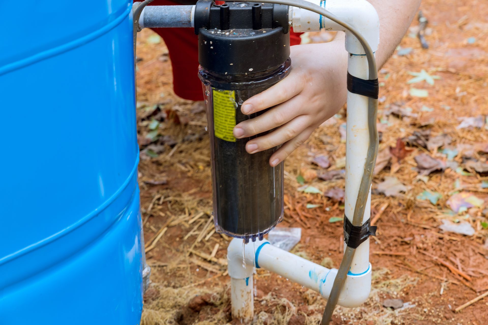 Hand adjusting a black water filter attached to white pipes beside a blue tank outdoors