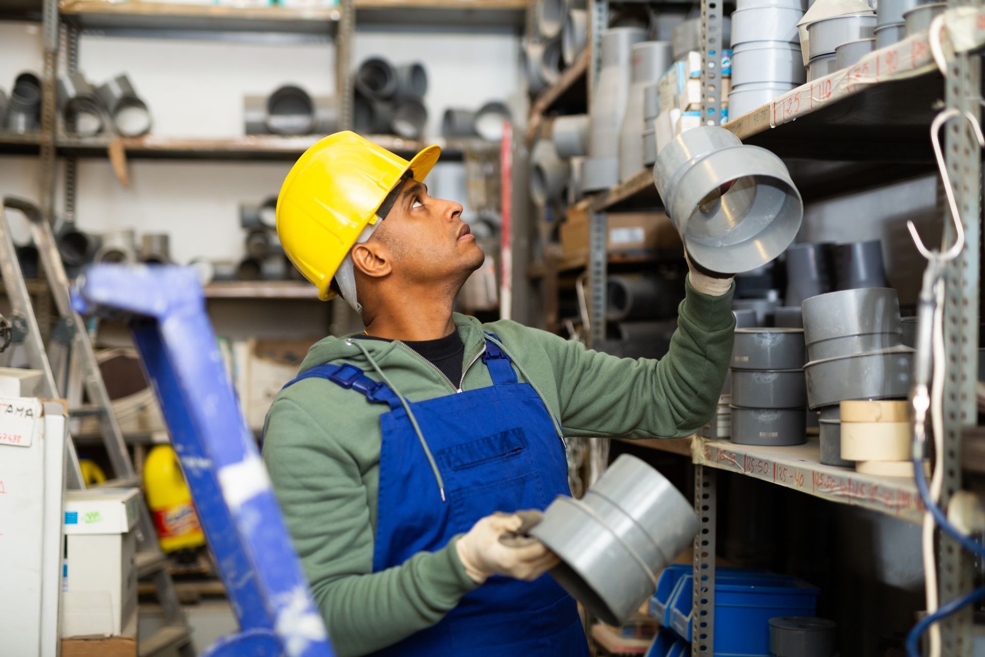 Worker in yellow hard hat inspecting metal ducting on a warehouse shelf