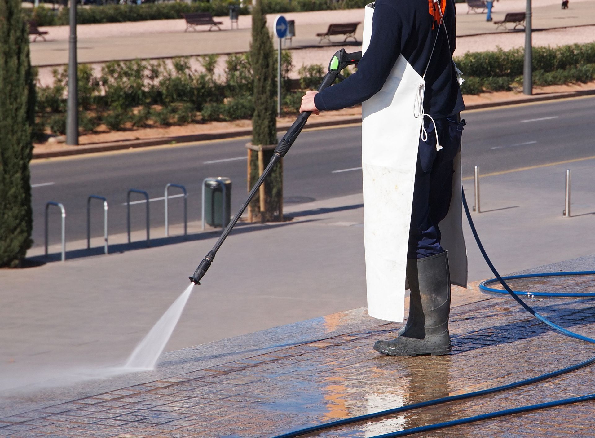 Worker pressure-washing a sidewalk with a long spray wand beside a road
