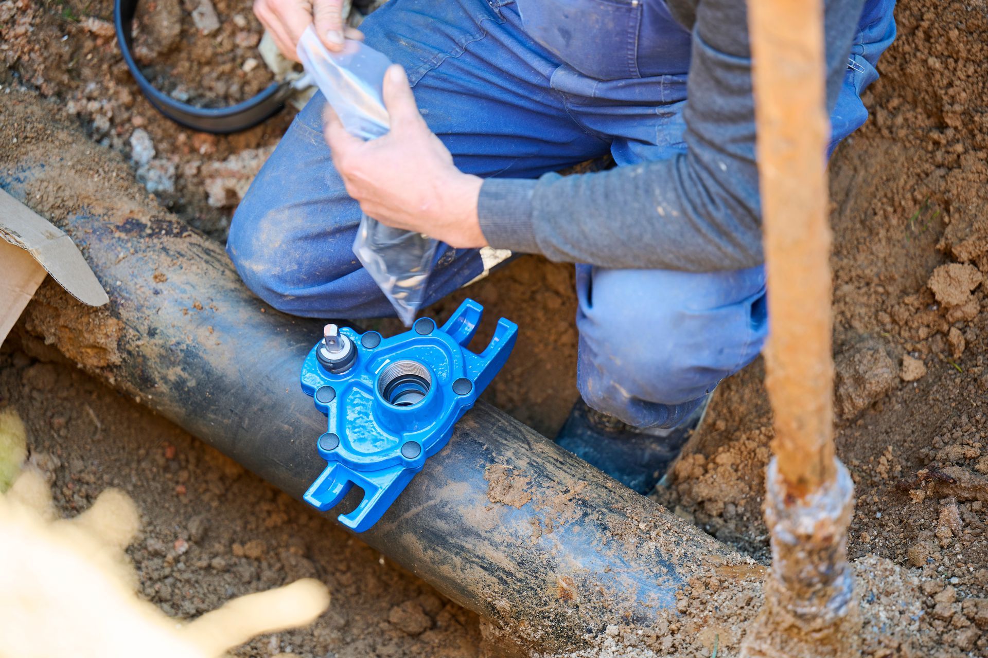 Worker kneeling in a trench, fitting a blue pipe valve on muddy ground.