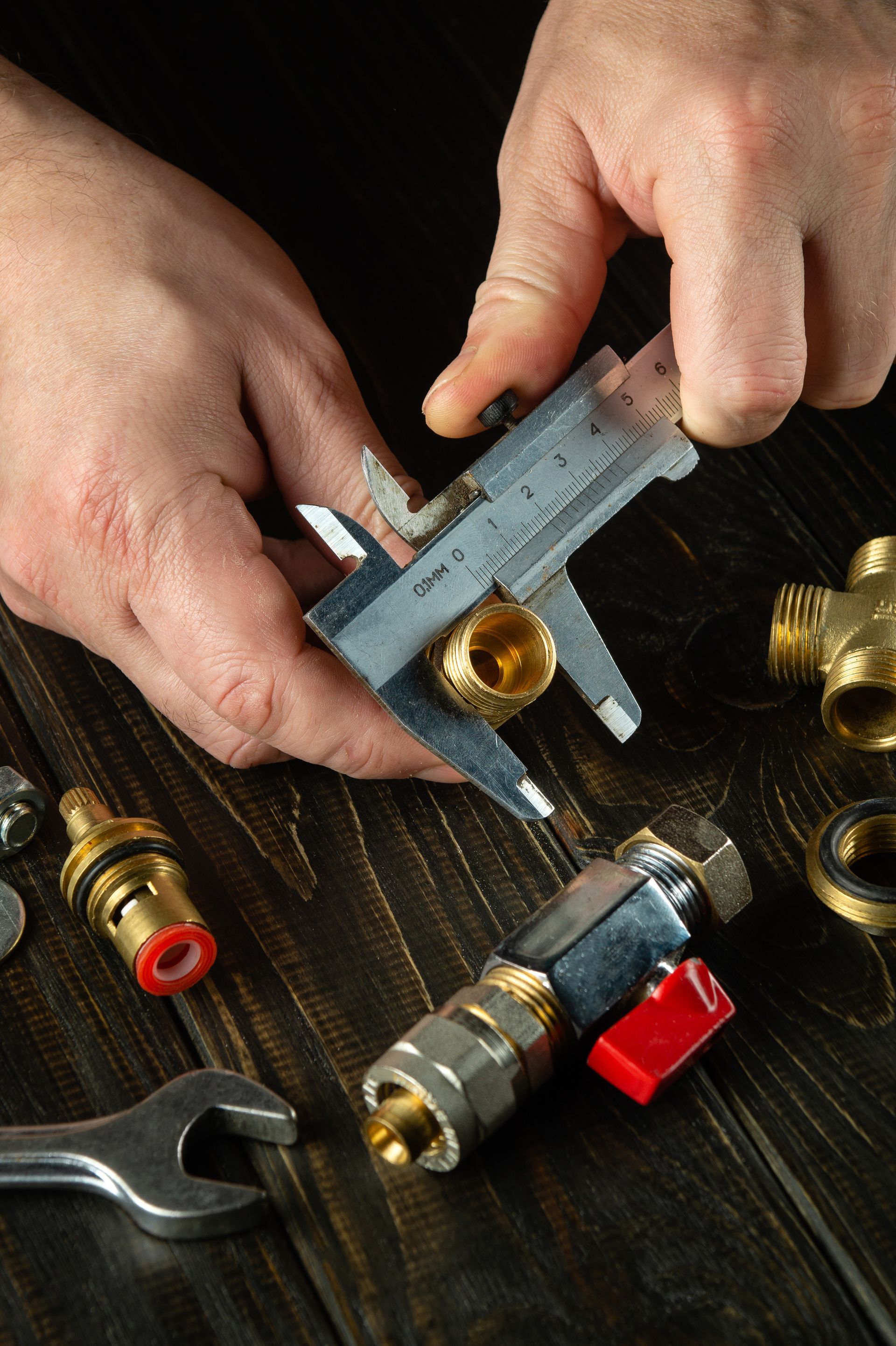 Hands using a caliper to measure a brass pipe fitting on a dark workbench.
