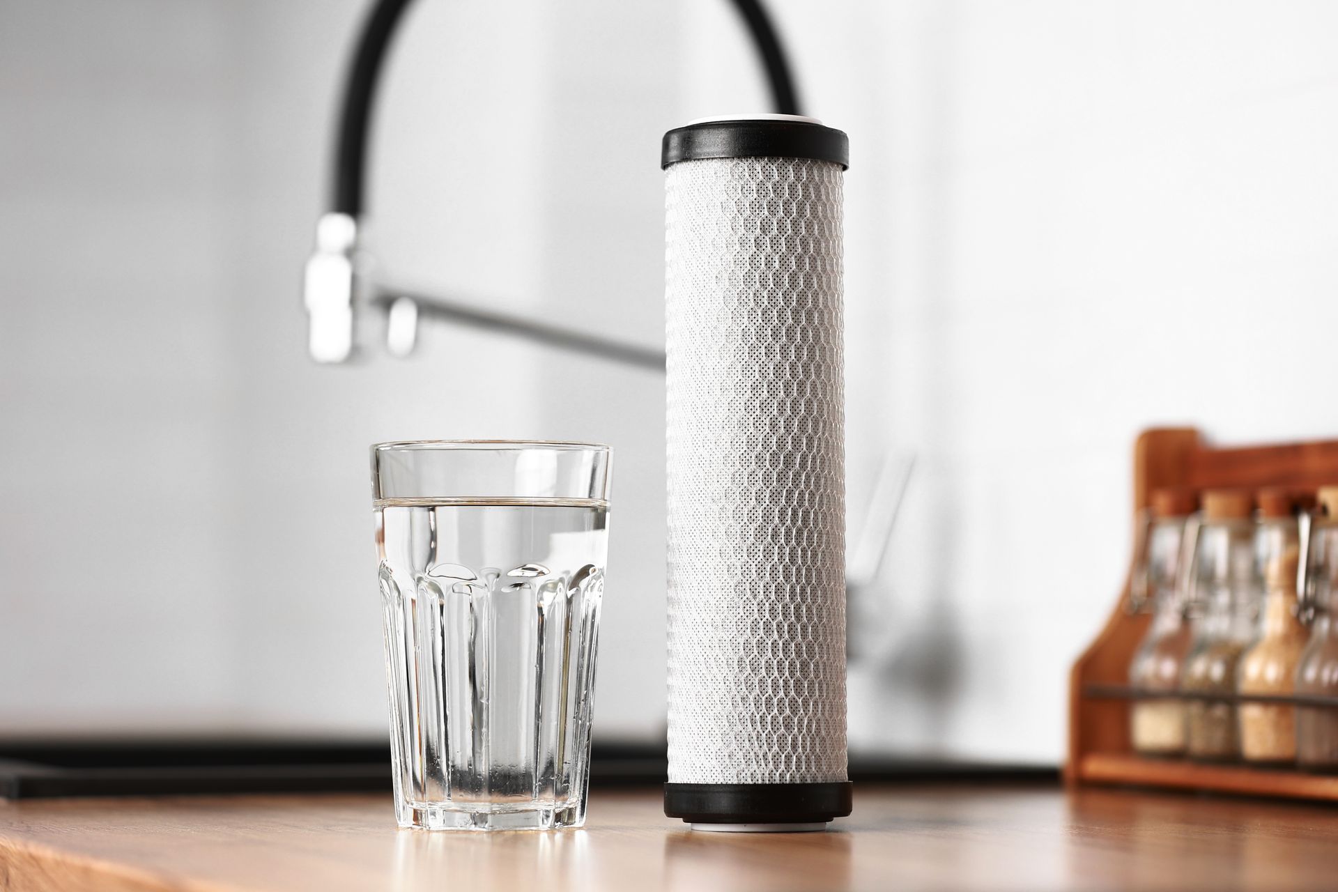 Glass of water beside a white textured travel bottle on a wooden table, with a faucet in the background