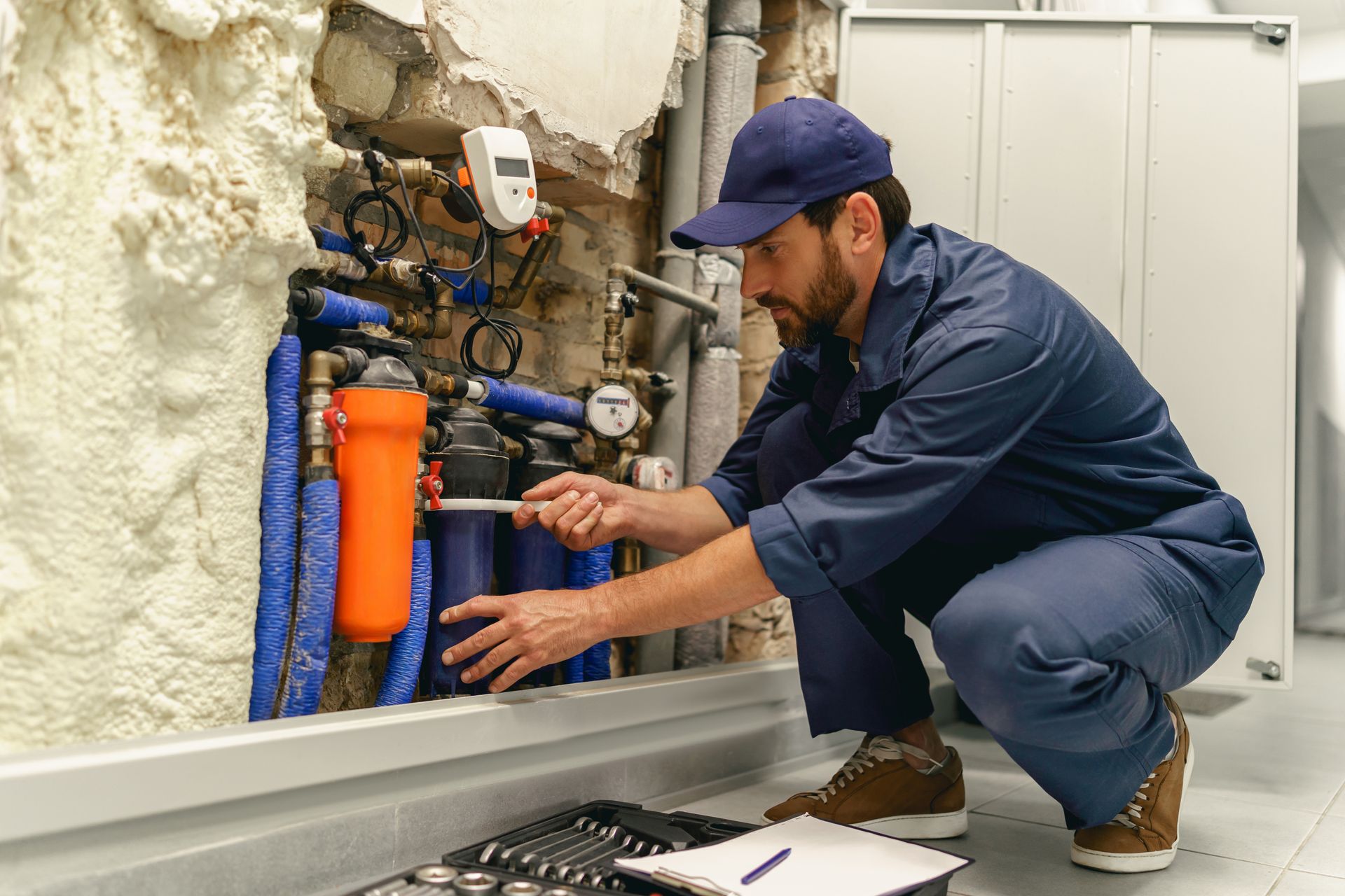 Plumber crouches beside exposed pipes, inspecting a wall-mounted plumbing system in a utility room.