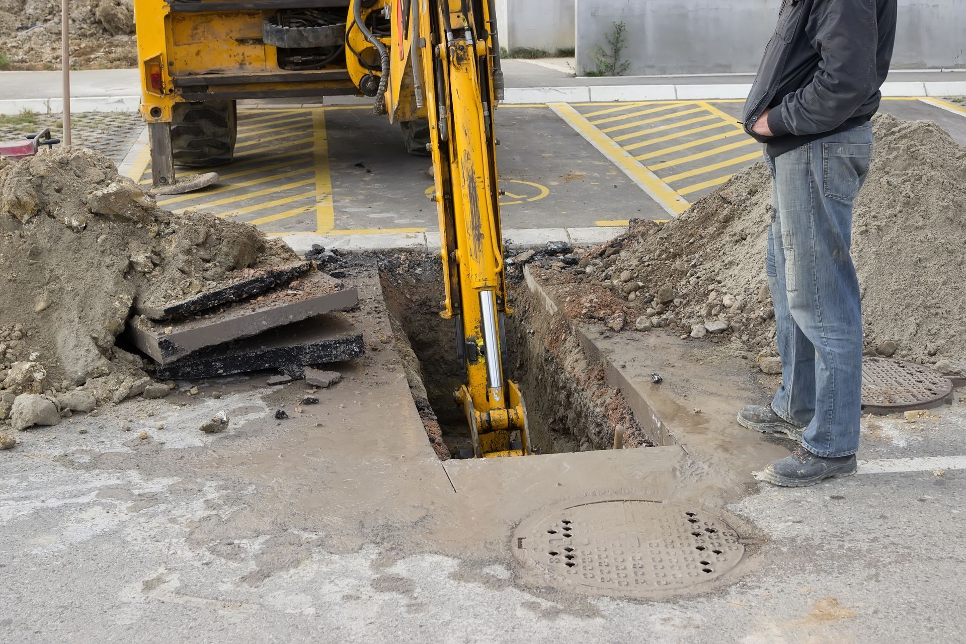 Yellow excavator digging a trench in a street, with a worker standing nearby