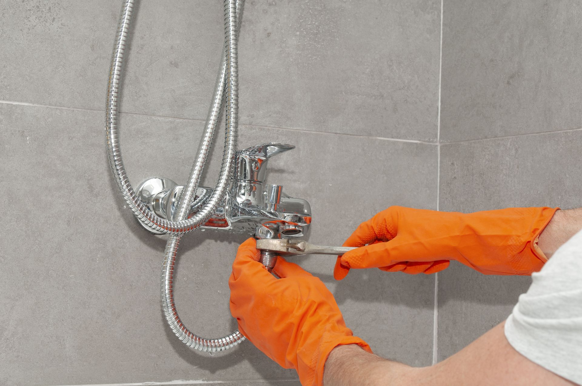 Person in orange gloves adjusting a chrome shower faucet on a tiled bathroom wall.