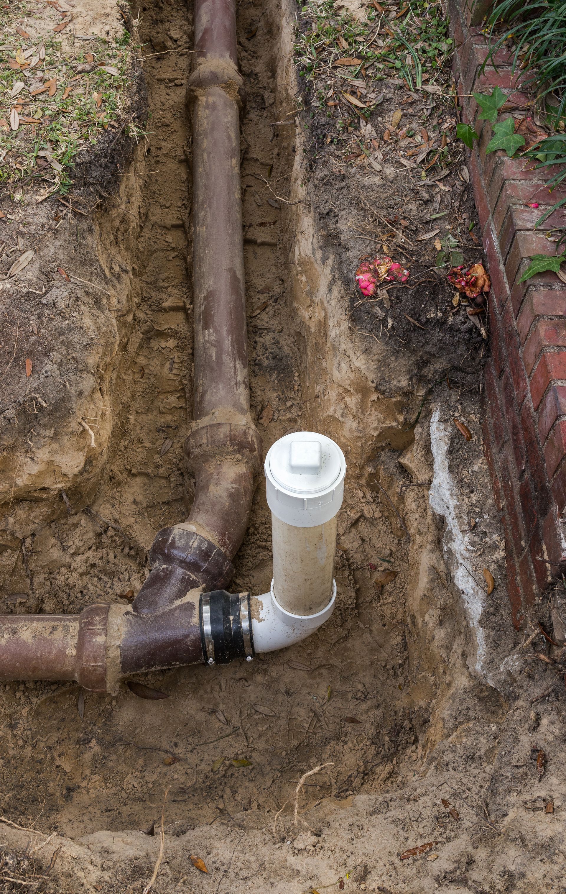 Underground plumbing with exposed white PVC pipe and elbow joint in a dirt trench beside a brick wall