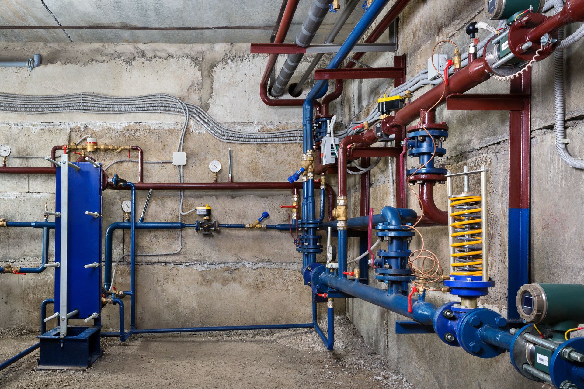 Industrial basement with blue and red pipes, valves, and tanks against a concrete wall.