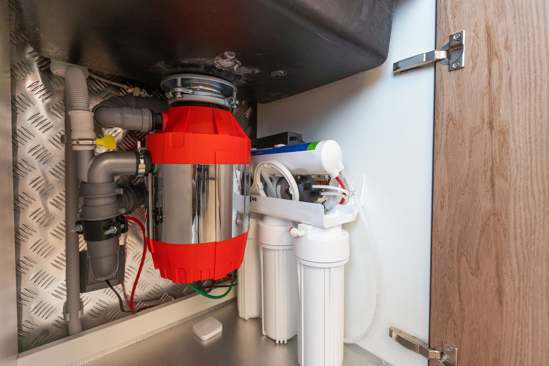 Under-sink plumbing with red garbage disposal and white pipes in a cabinet