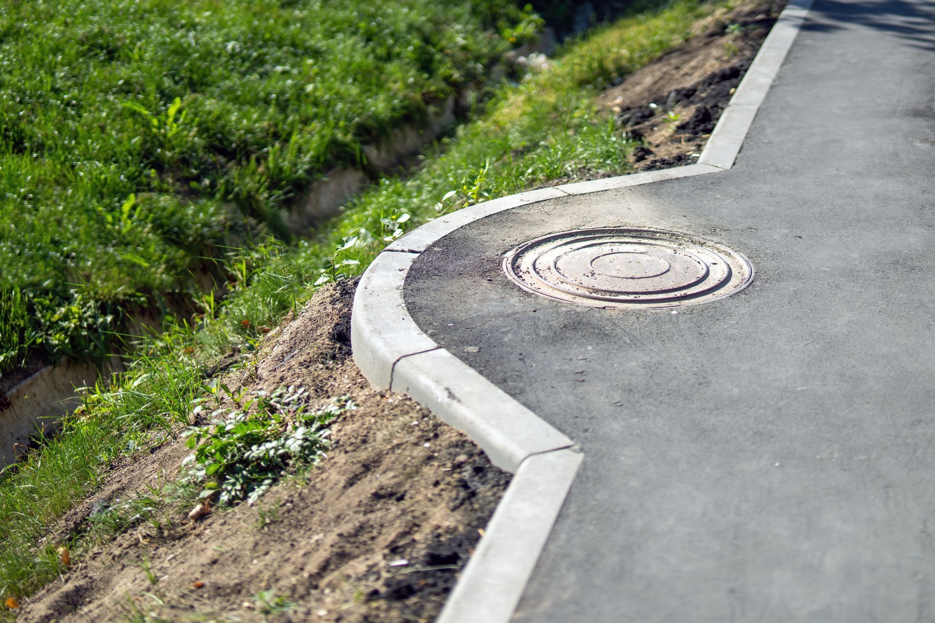 Curving roadside curb with a manhole cover beside a grassy slope and paved path