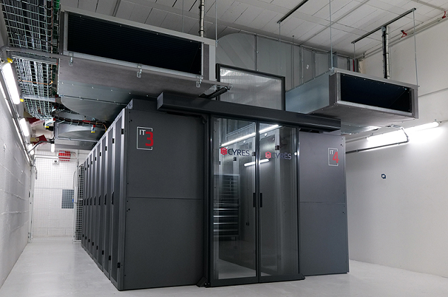 Un système de baies de serveurs sombres et fermées, avec des portes vitrées, installé dans une salle serveur industrielle lumineuse équipée de conduits d'aération.