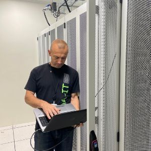 Une personne vêtue d'un t-shirt foncé utilise un ordinateur portable, debout dans une salle des serveurs à côté d'une rangée de baies de serveurs métalliques.