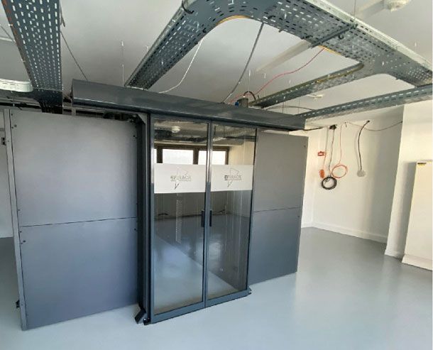 Une armoire serveur centrale en verre, entourée de panneaux métalliques, dans une pièce équipée de chemins de câbles suspendus.
