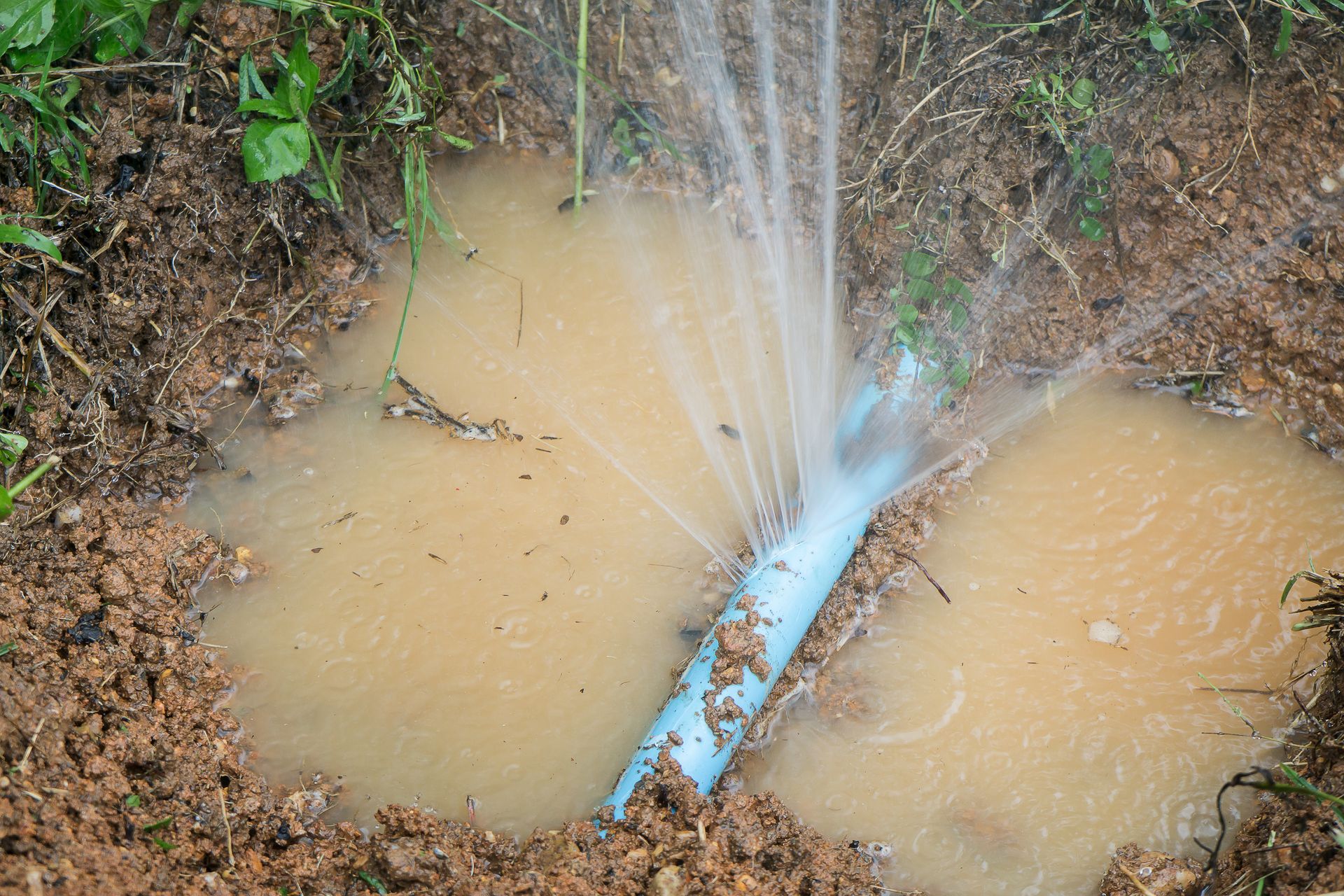 Water spraying from a broken blue pipe into muddy puddles in a dirt ditch