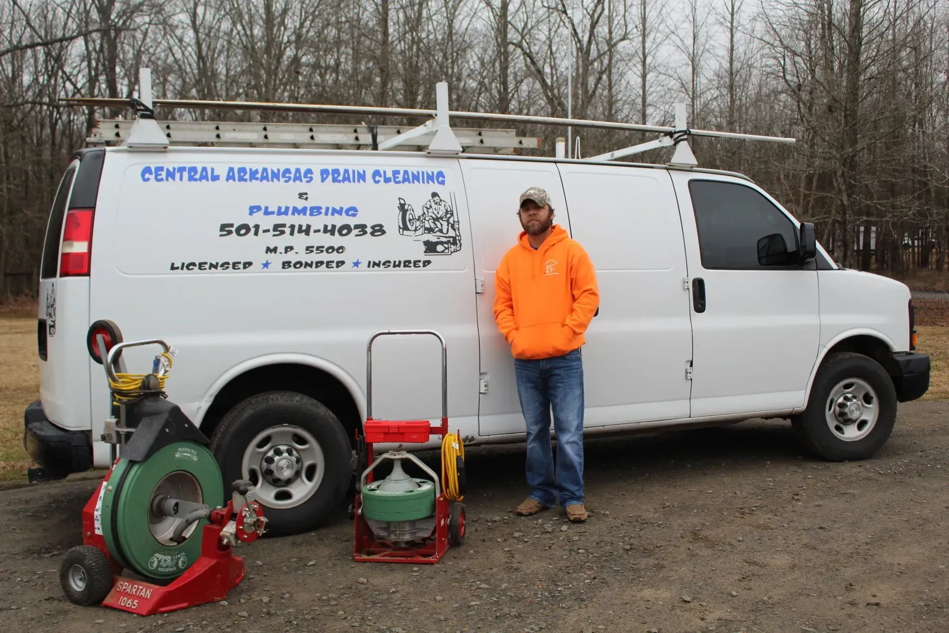 Worker in orange jacket beside white service van and sewer-cleaning equipment outdoors