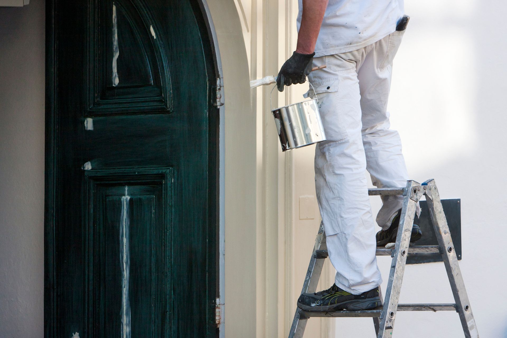 Painter on a ladder painting a white wall beside a dark green door