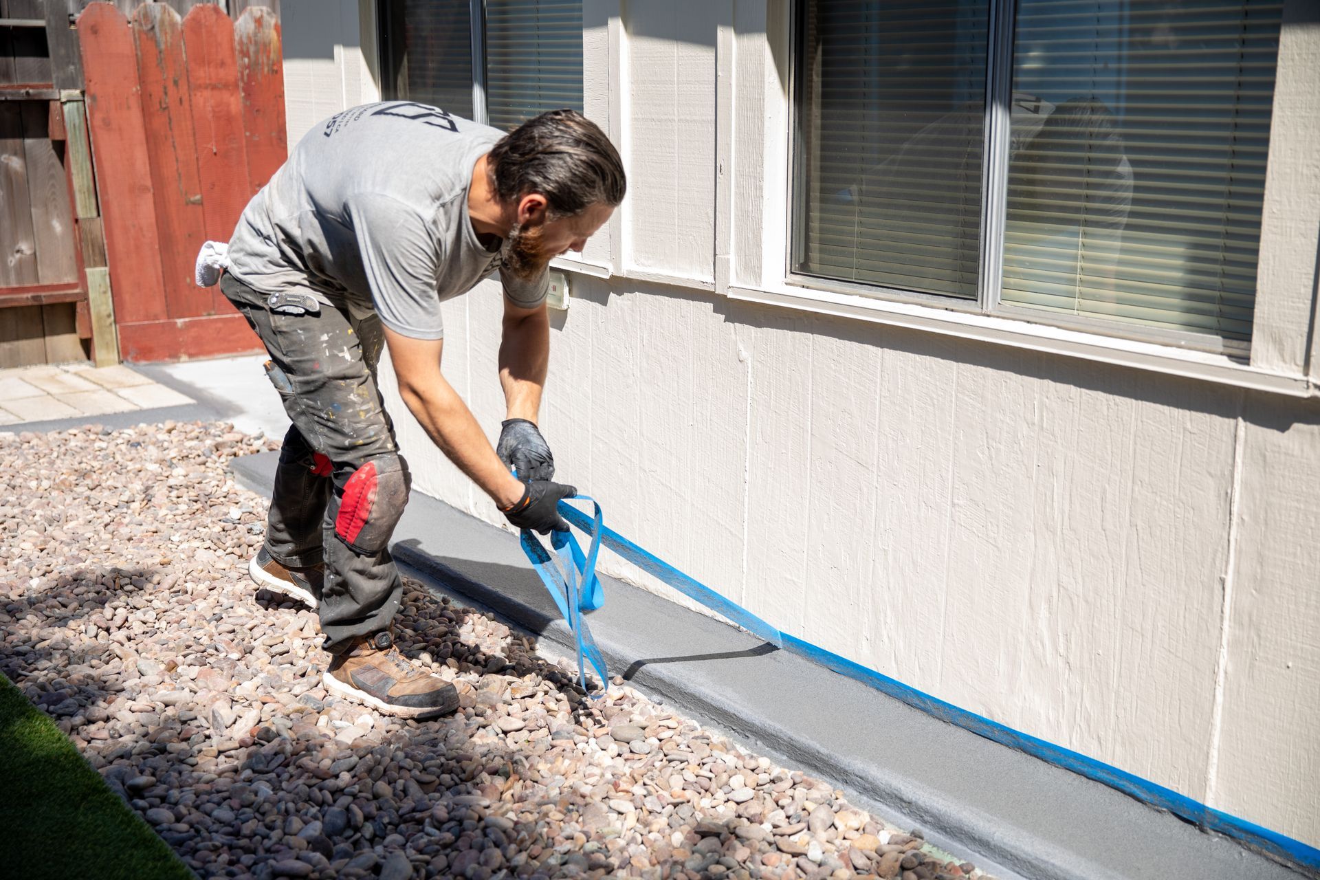 Worker applying blue tape along the base of a white house exterior wall