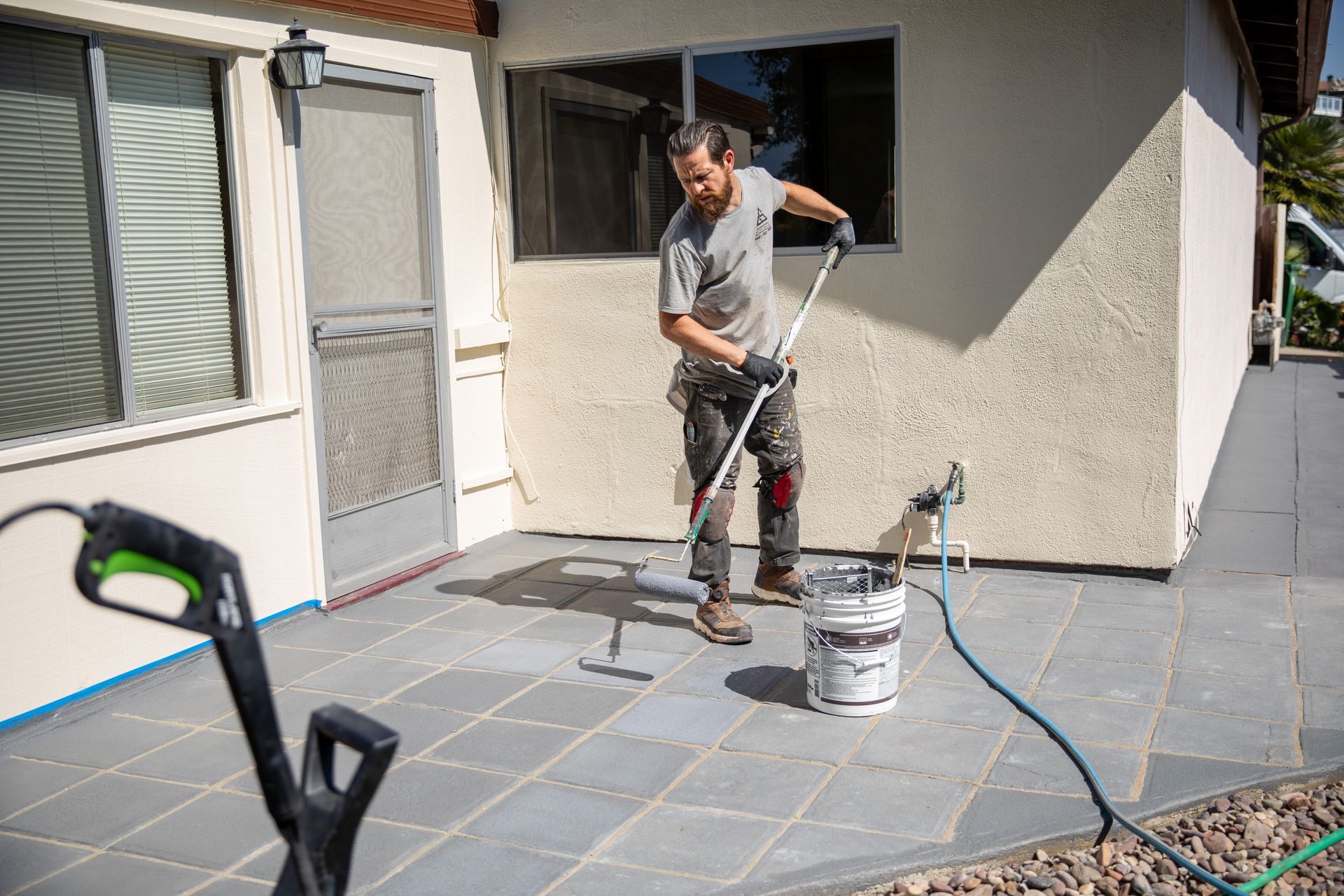 Person painting a house exterior patio with a roller beside a paint bucket and hose