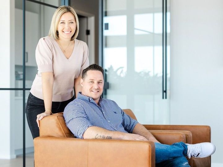 Woman leaning on man seated in brown leather chair, smiling. Glass office setting.
