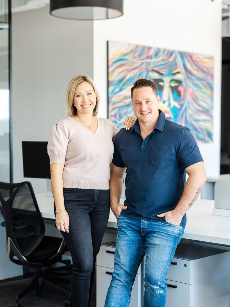 Woman and man pose in office; woman in light top and dark jeans, man in blue shirt and jeans. Painting of lion behind them.