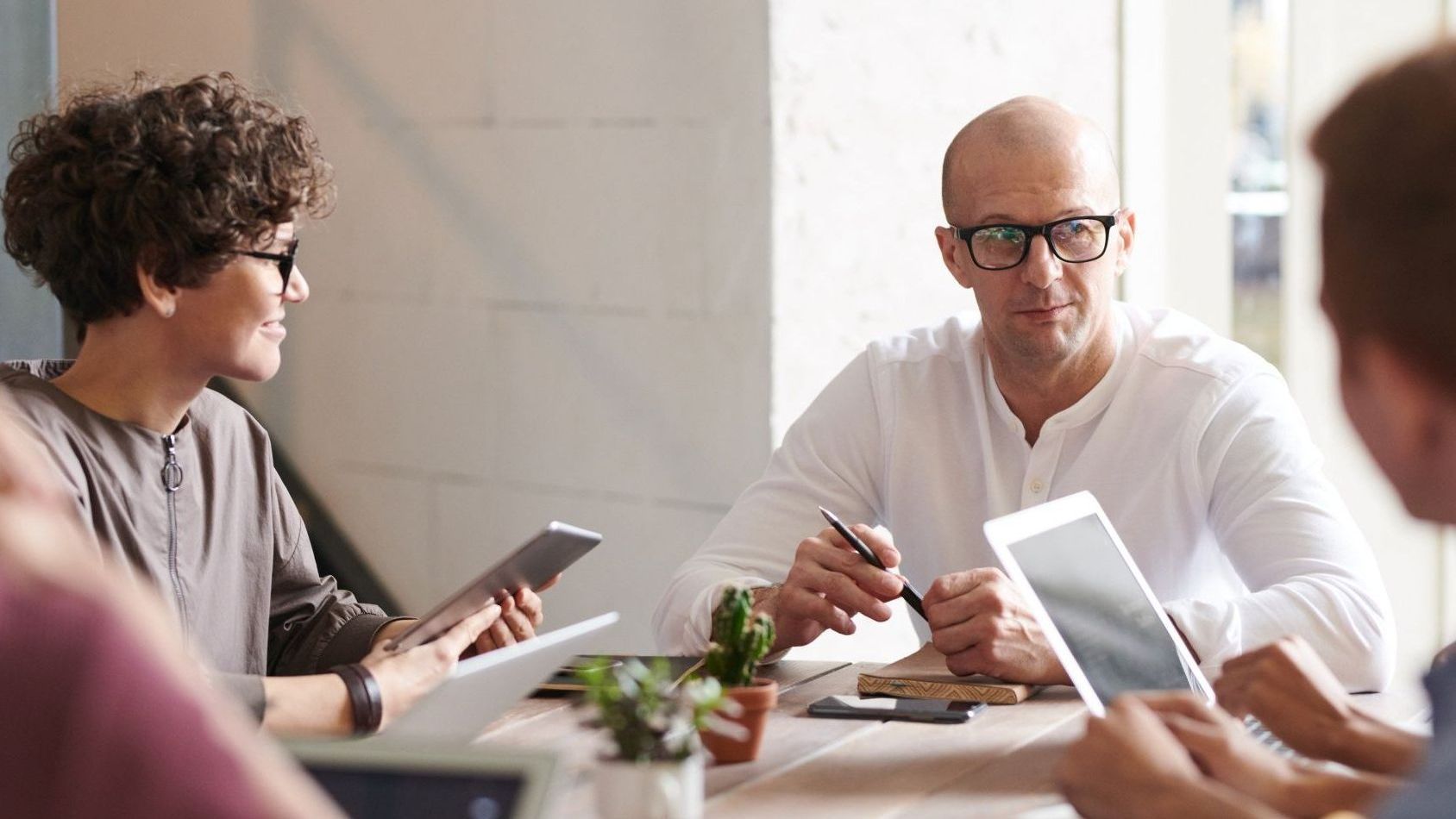 People at a table, some using tablets. A bald man with glasses looks intently; another person smiles. Modern office.