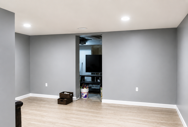 Empty gray room with light wood flooring, recessed lights, and an open doorway to a cluttered adjoining room