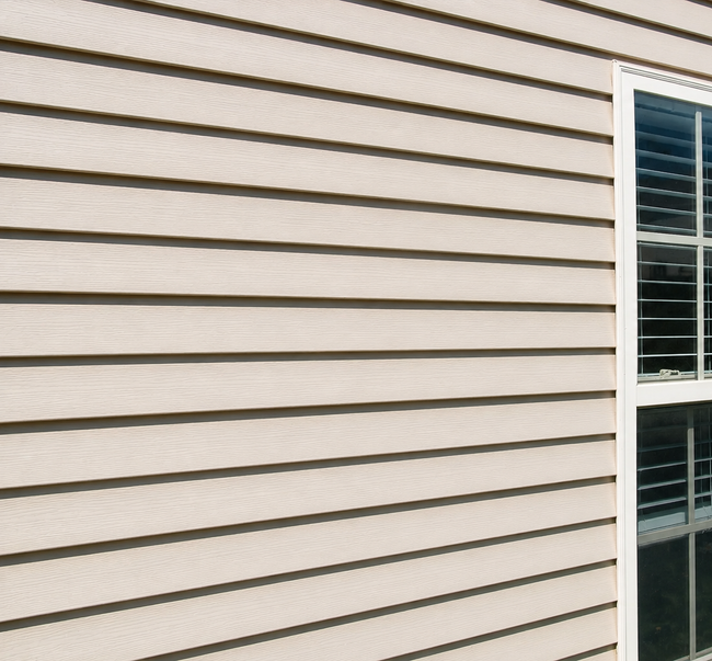Beige house siding beside a white-framed window with closed blinds and greenery below