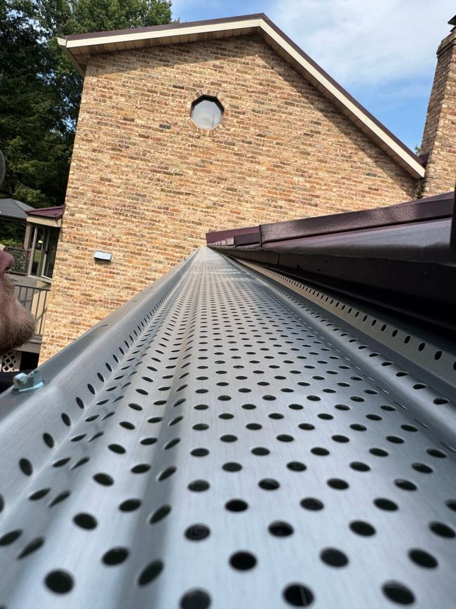 Perforated metal roof gutter leading to a brick house wall with a round window under a cloudy sky