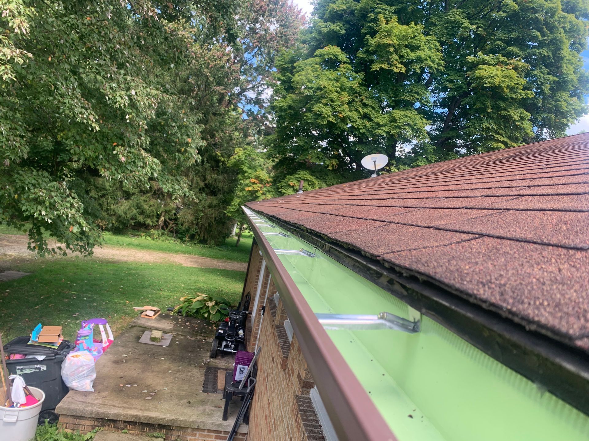 Roof edge with green gutter beside a wooded yard and patio items below