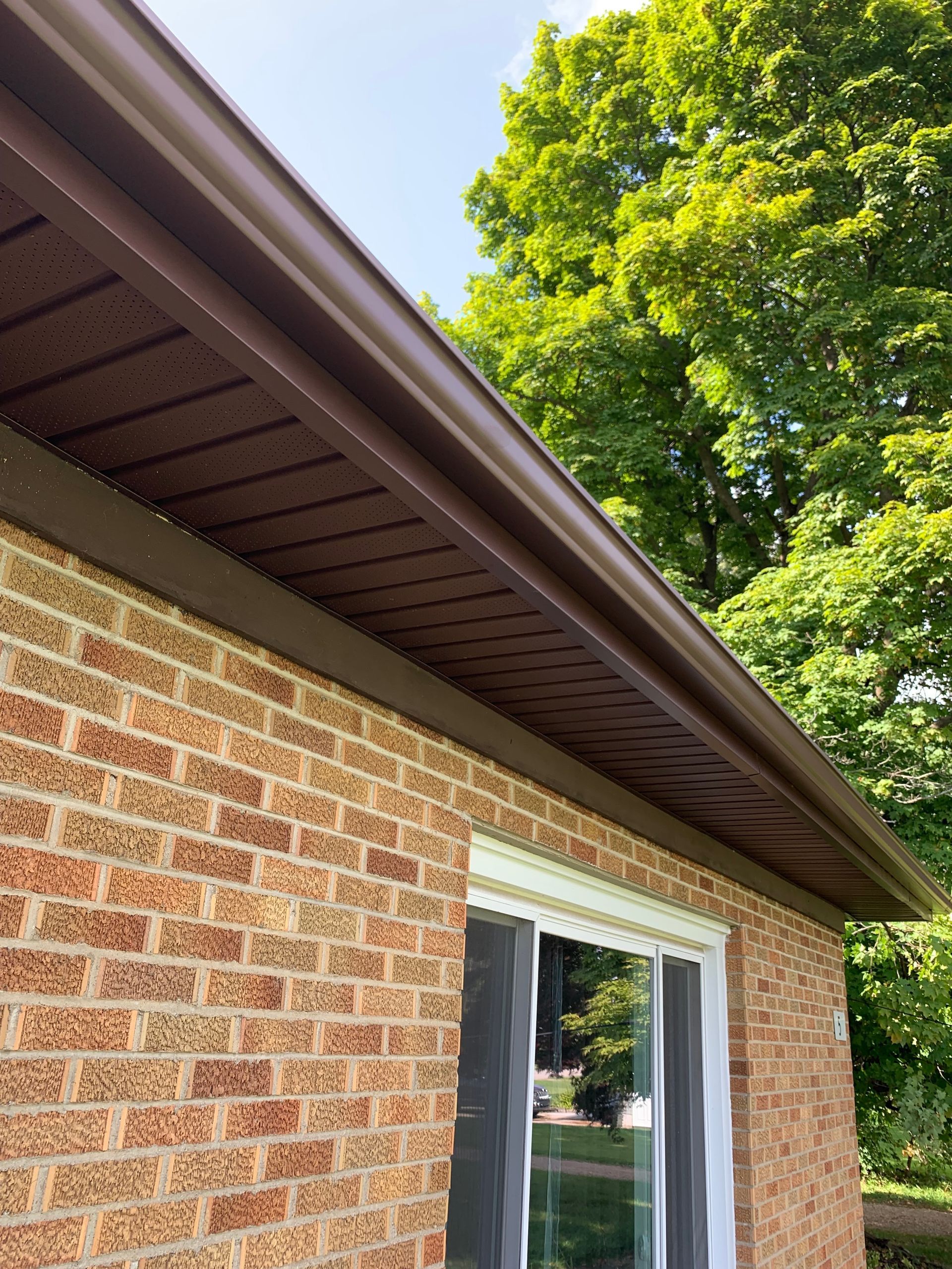 Brick house exterior with brown roof eaves and a white sliding window under a leafy green tree