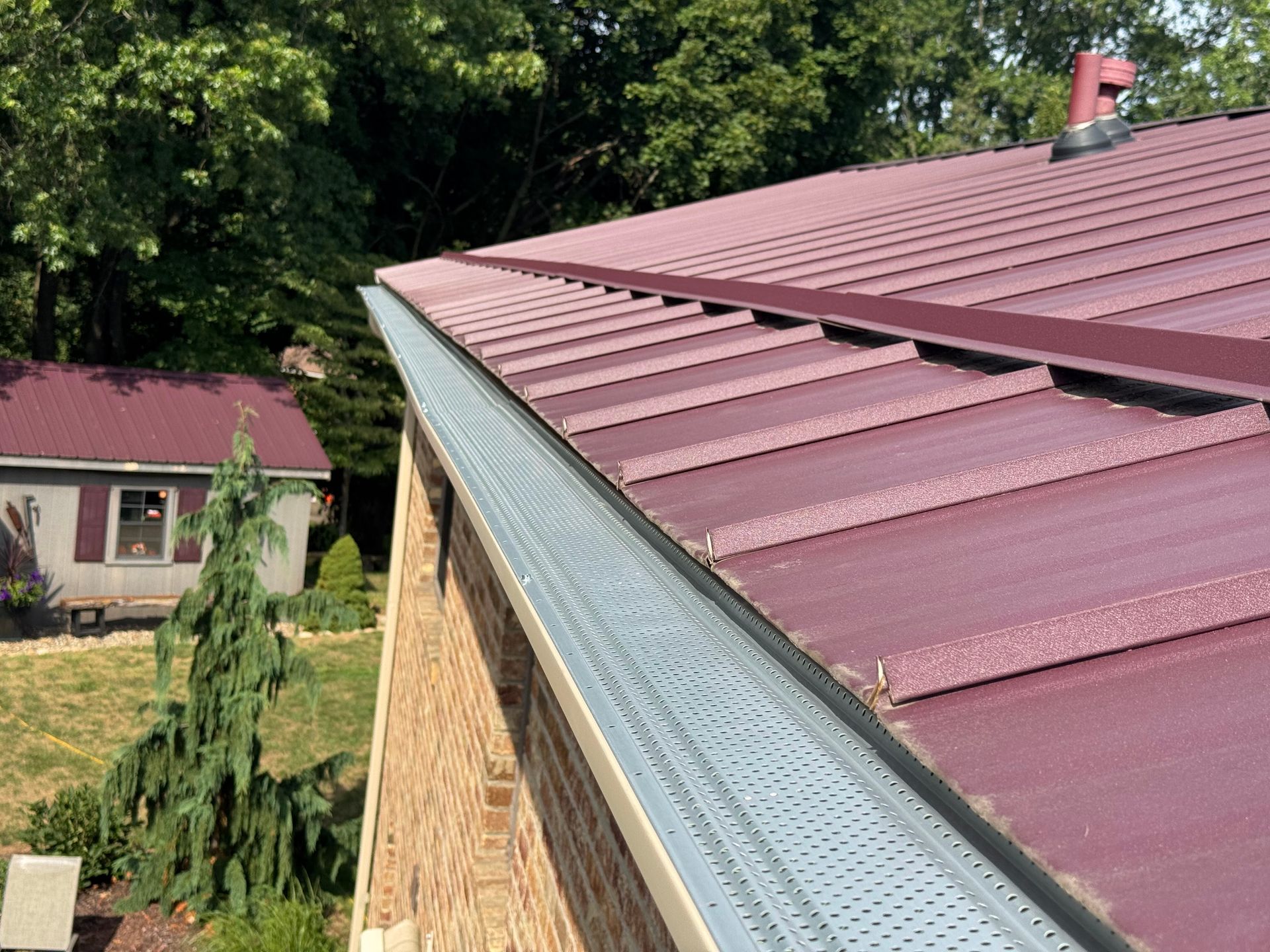 Red metal roof with gutter on a house, viewed from the eaves beside trees and a yard