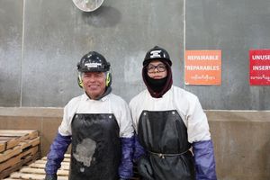 Two workers in white protective suits and black aprons standing indoors, smiling in front of a concrete wall.