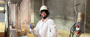 Worker in white protective clothing and hard hat standing in an industrial room, holding a blue device.