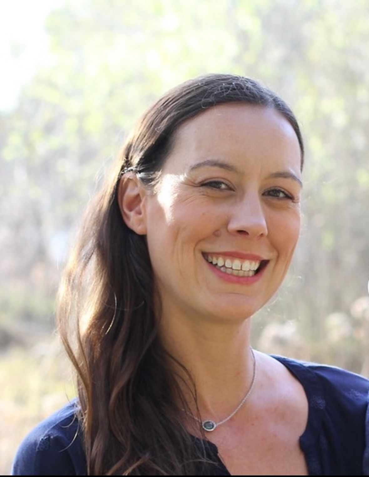 A woman wearing a blue shirt and a necklace is smiling for the camera.