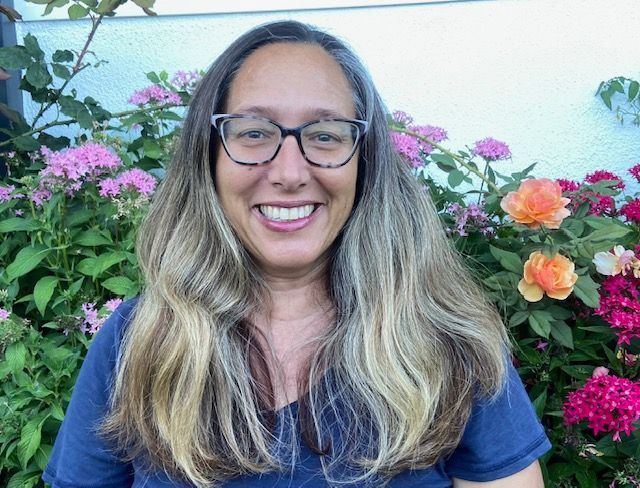 Smiling person with glasses in front of pink and orange flowers outdoors