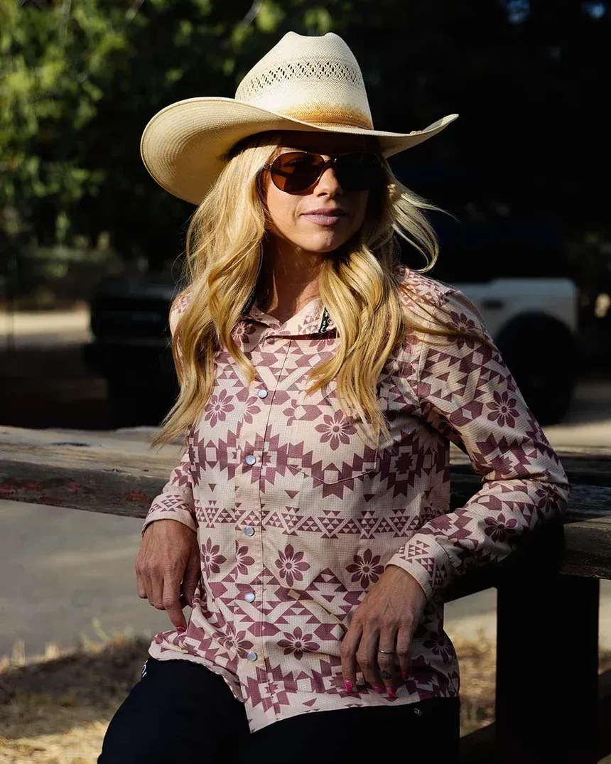 Woman smiling, holding daisy over eye, wearing floral shirt and hat, outdoors.