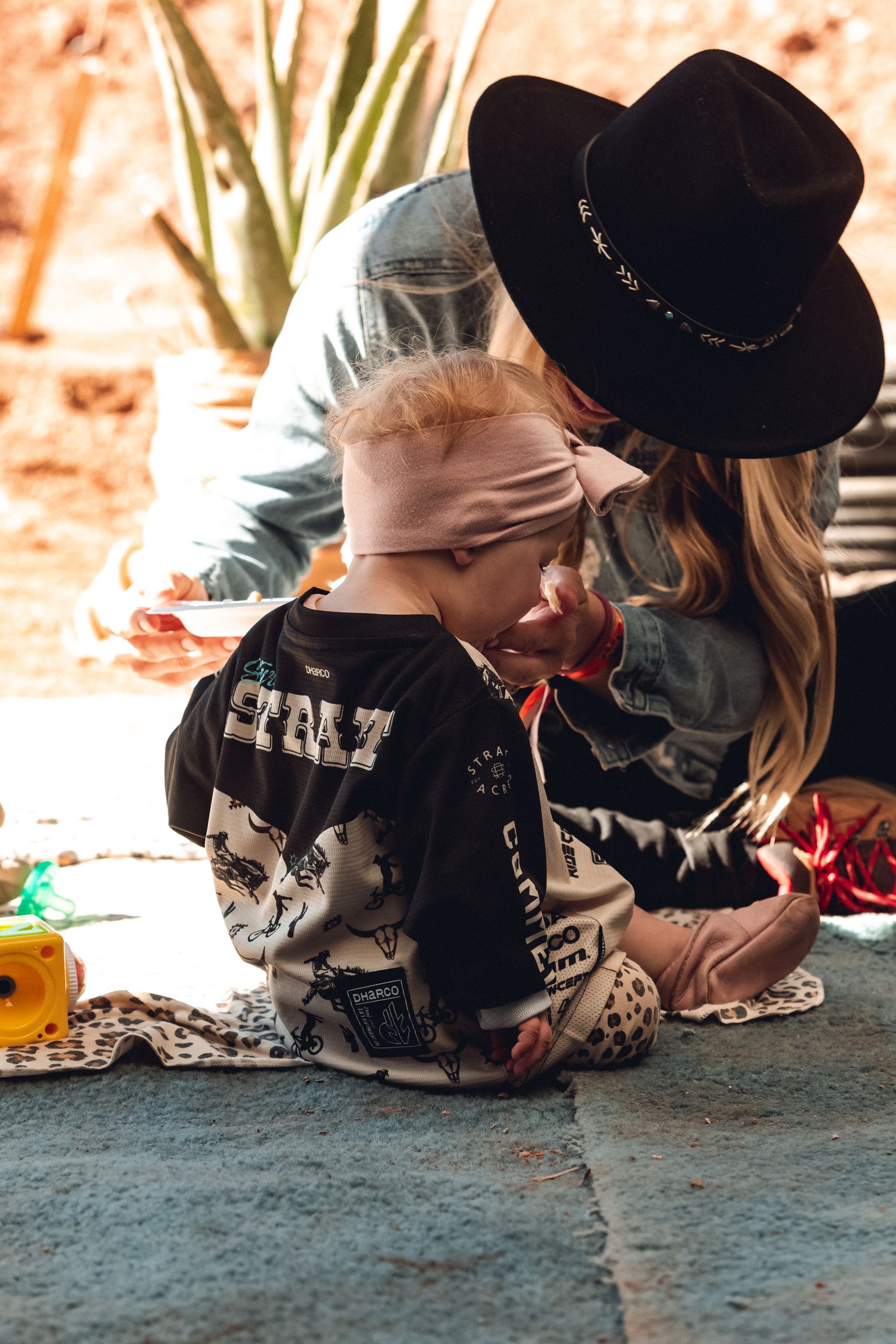 A young child with a pink headband sits on a blue surface, playing while an adult wearing a black hat looks on.