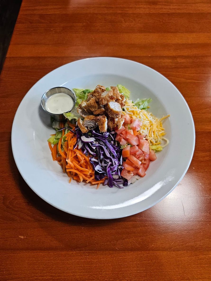 Salad with fried chicken, shredded carrots, cabbage, cheese, and tomatoes; side of white dressing on a white plate.