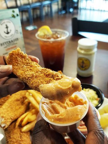Person dipping fried fish into a small container of orange dipping sauce, with fries and a beverage in the background.