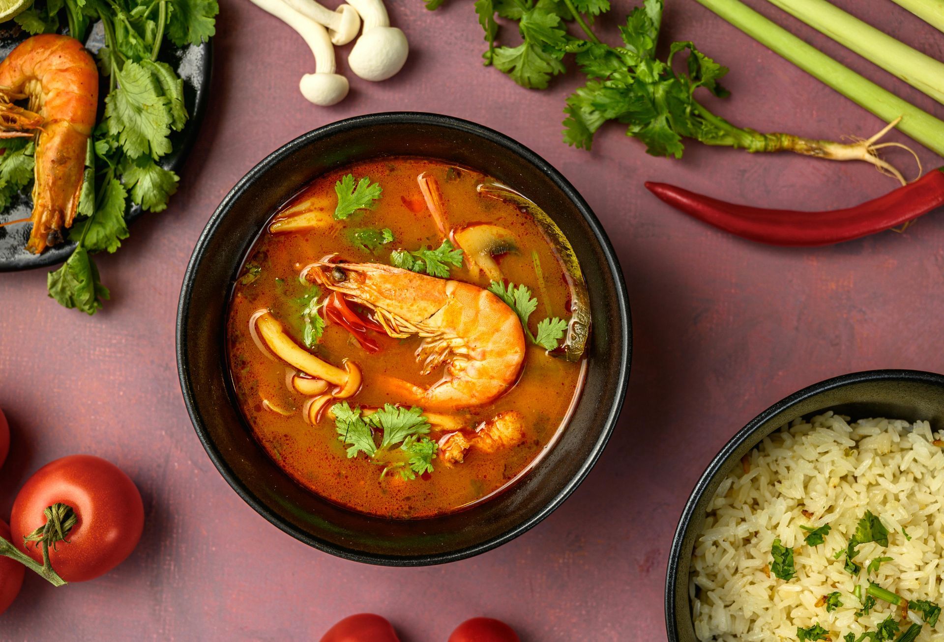 A bowl of Tom Yum soup with shrimp, surrounded by ingredients like rice, tomatoes, mushrooms, and herbs on a purple table.