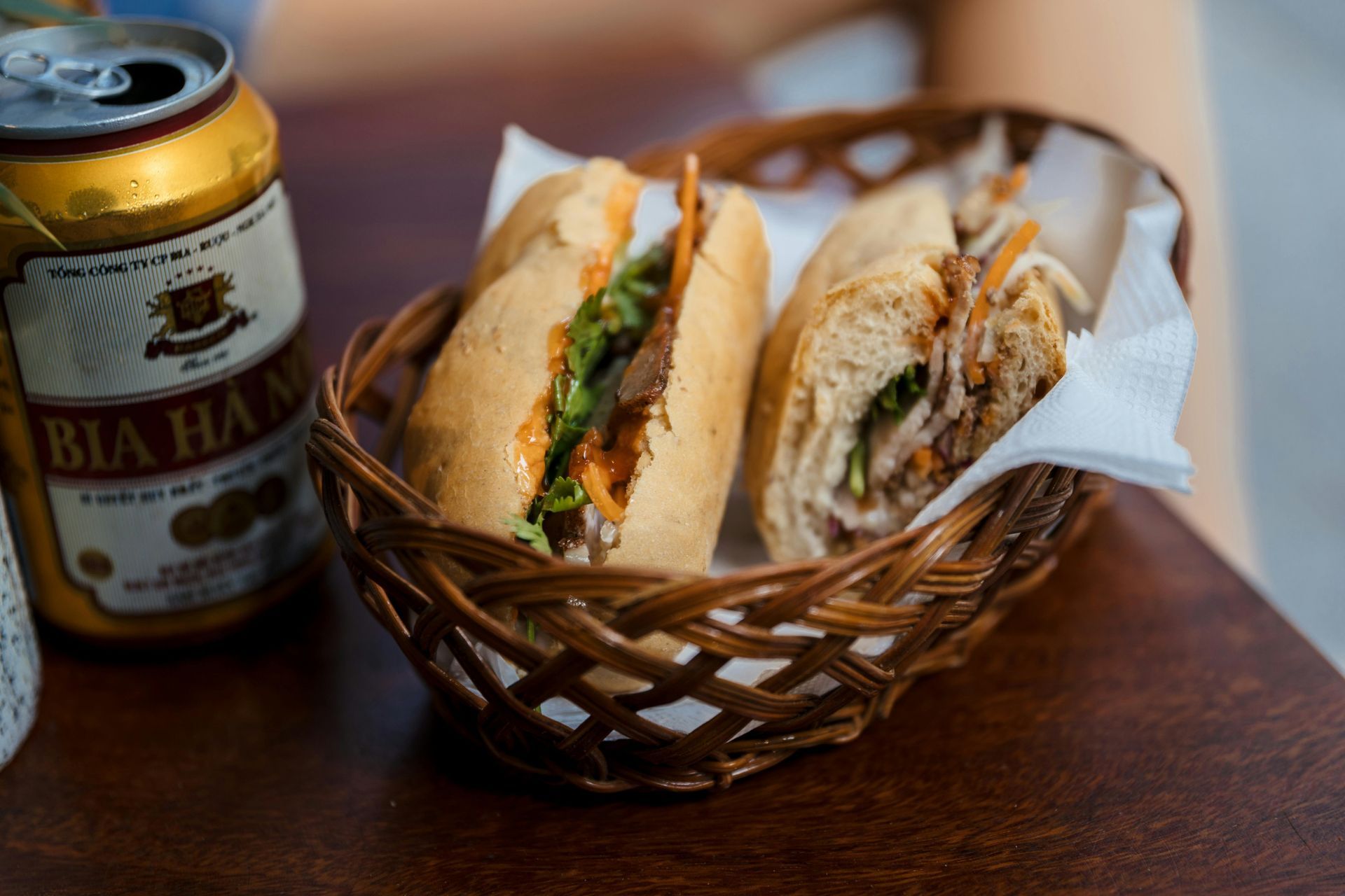 Two sandwich halves in a wicker basket beside a beer can on a wooden table