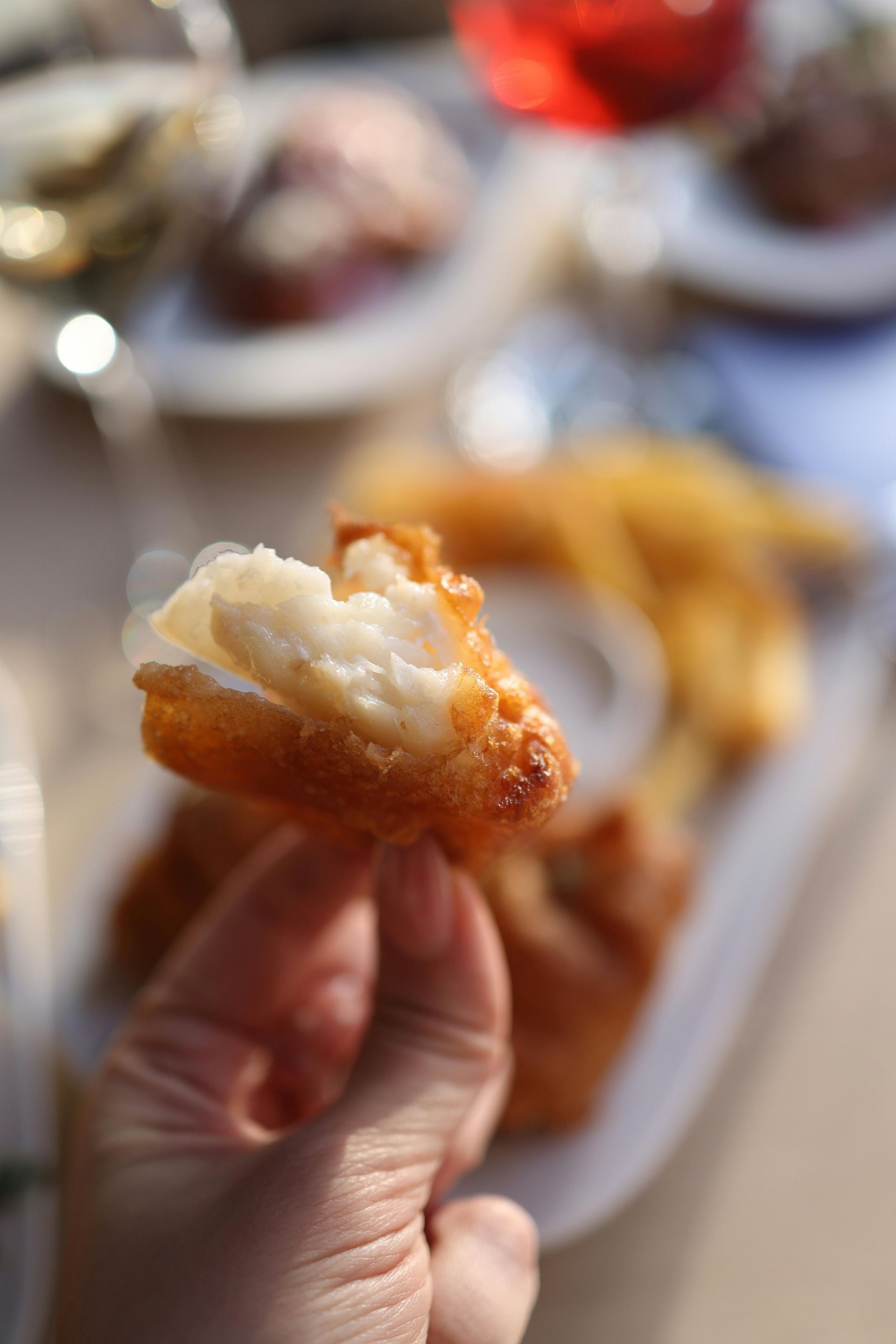 Hand holding a fried, golden-brown food item with a creamy, white interior, other plates in the blurred background.