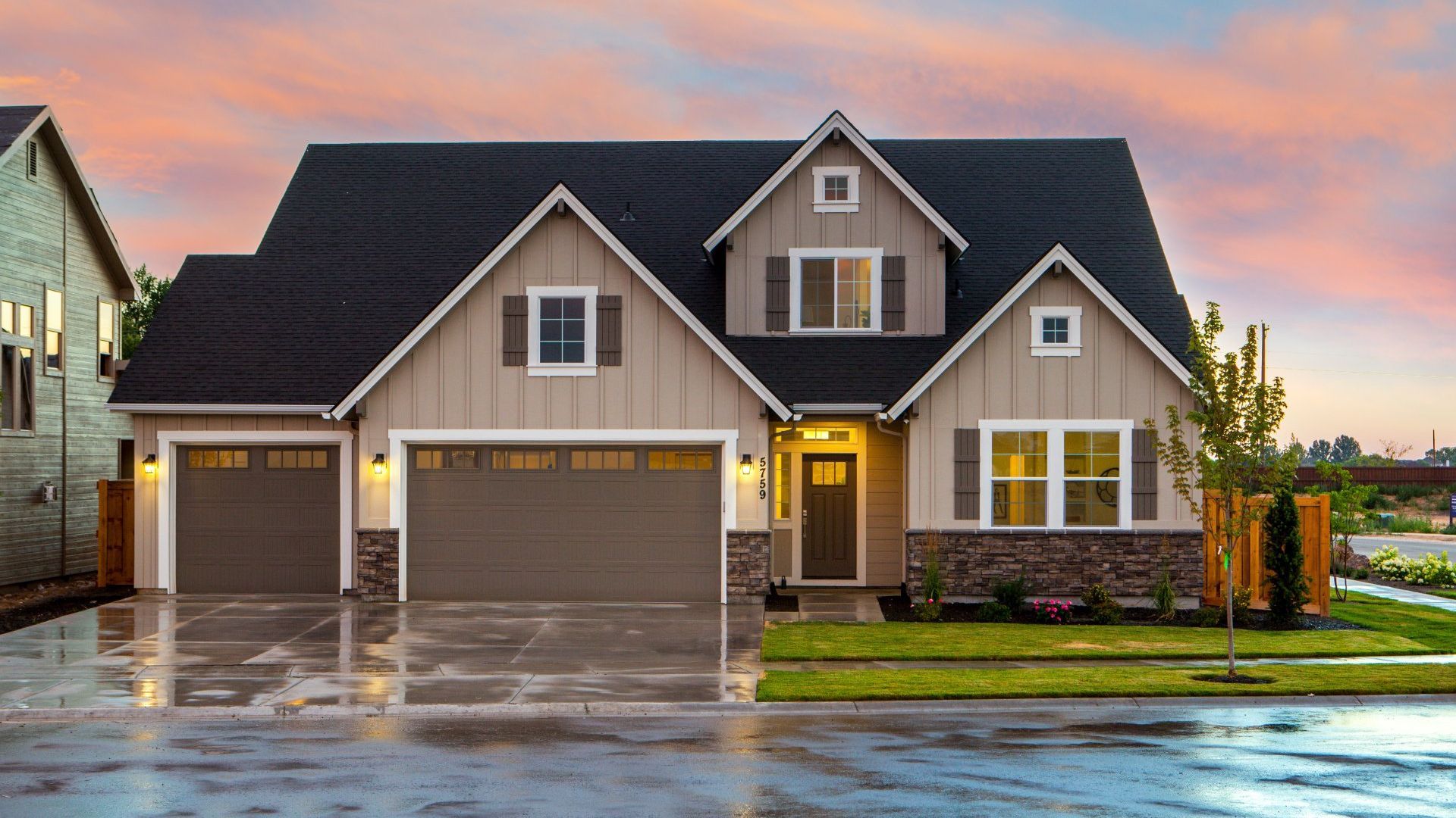 Suburban house with lit porch lights and three-car garage on a wet driveway at dusk