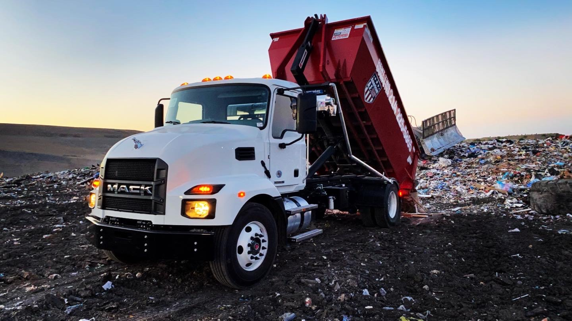 a white mack dump truck is parked in a landfill dumping a 20 yard dumpster full of waste.