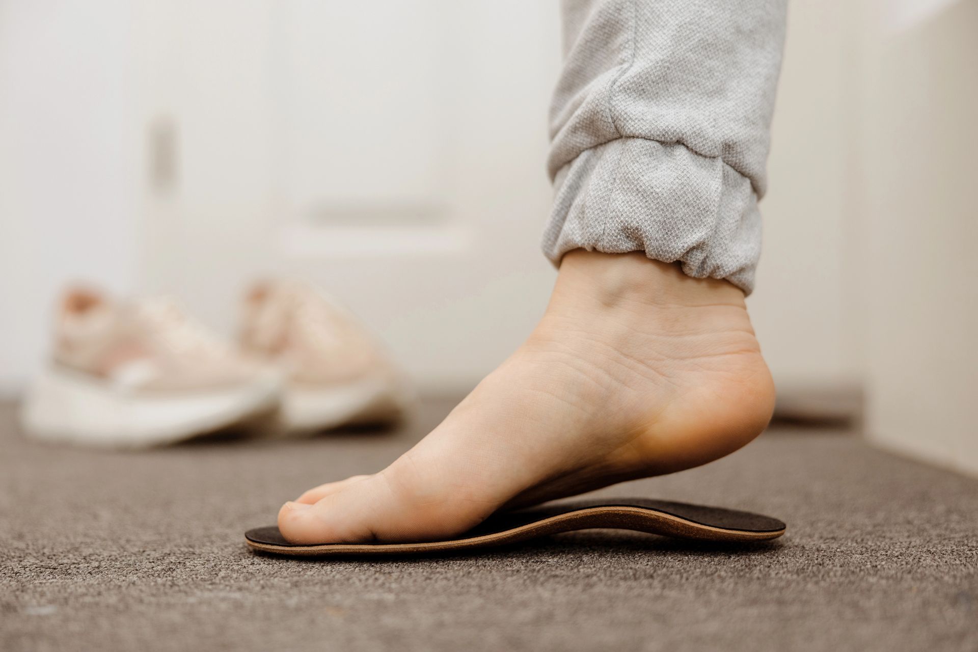 Il piede nudo di una persona appoggiato su una soletta sagomata su un pavimento ricoperto di moquette, con un paio di scarpe da ginnastica sullo sfondo.
