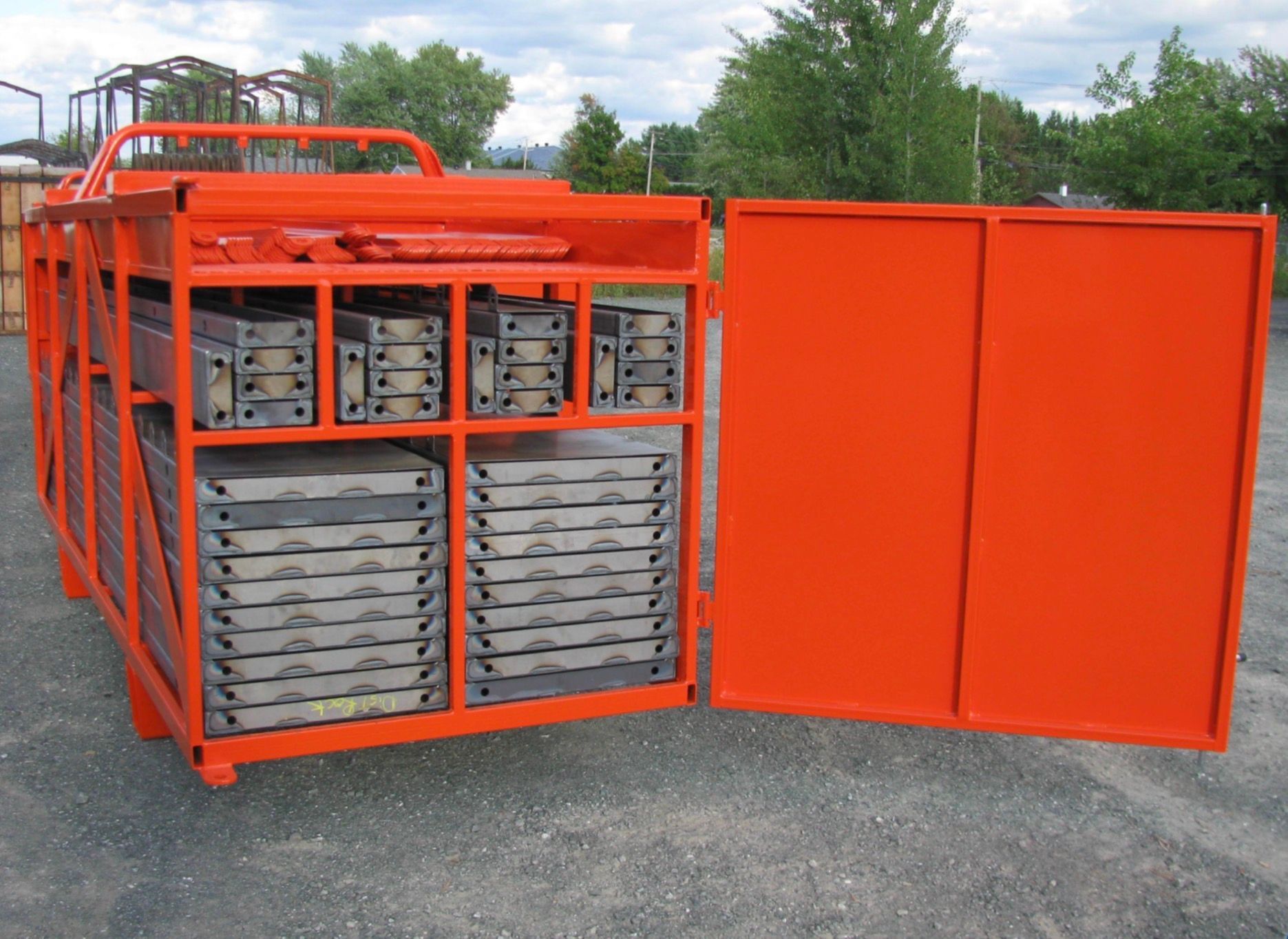 Étagères de rangement en métal orange avec plateaux gris empilés, installées à l'extérieur sur un terrain gravillonné.