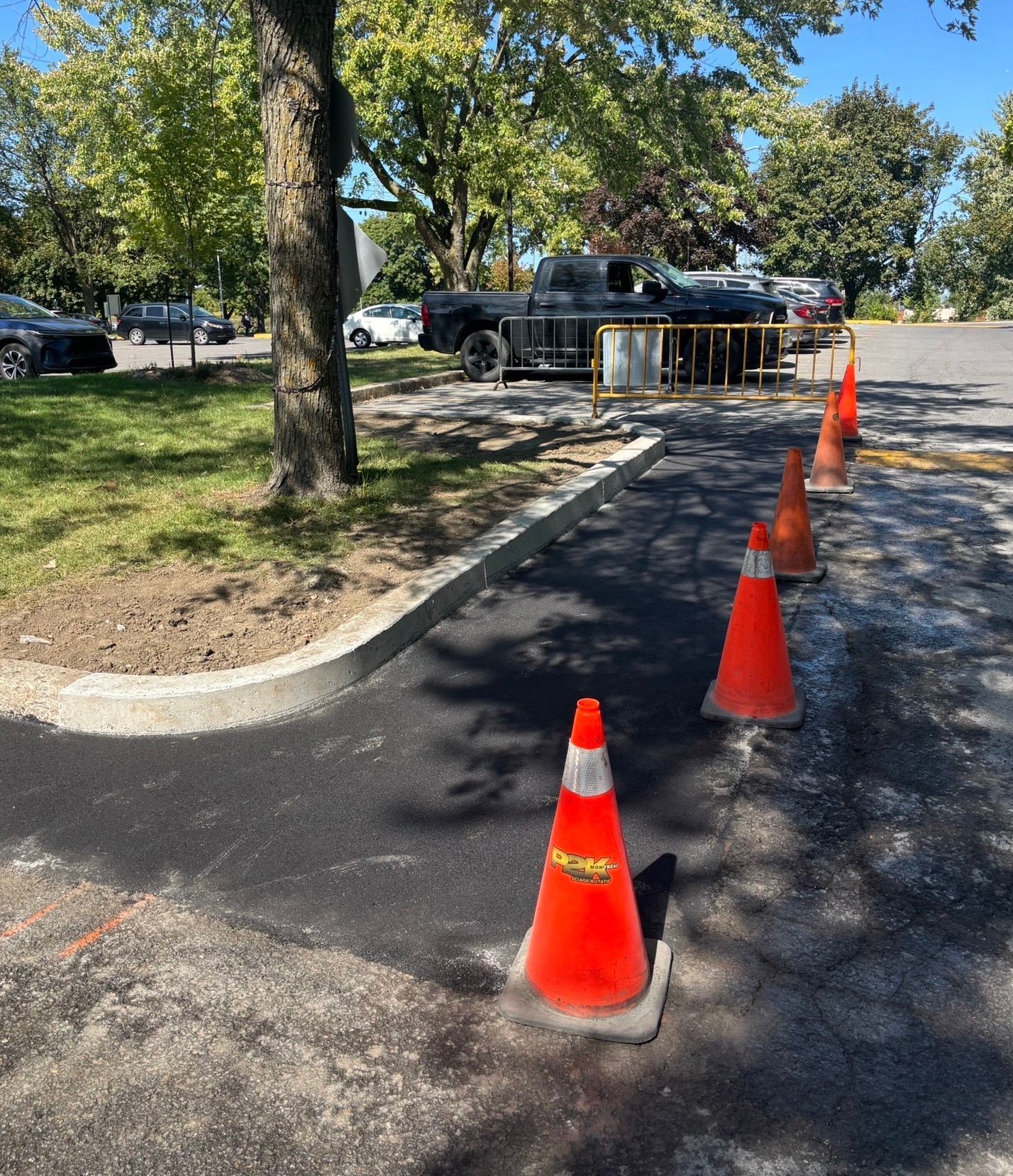 Des cônes de signalisation orange bordent une nouvelle rampe d'accès en asphalte noir dans un parking ensoleillé.