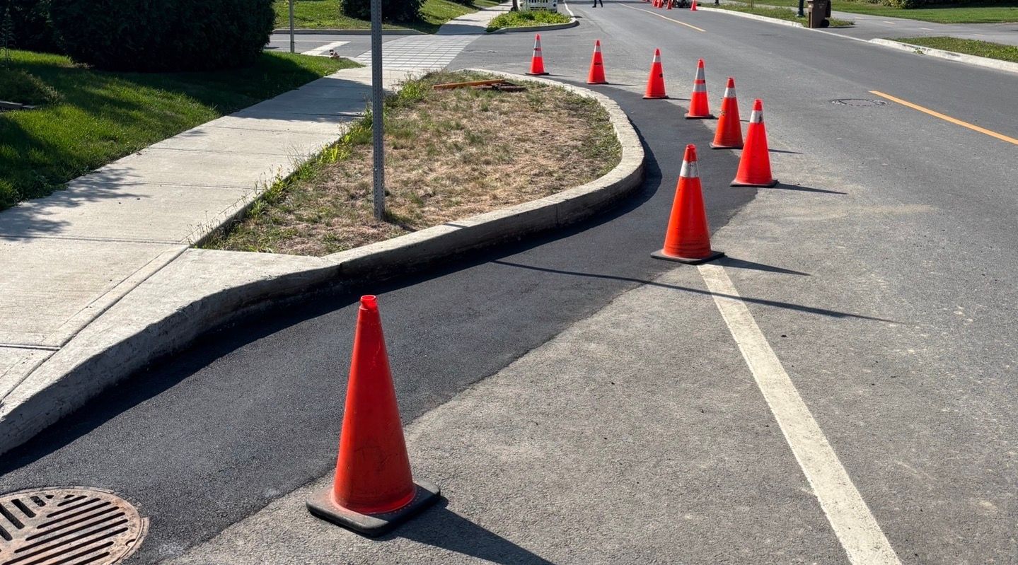Des cônes de signalisation orange bordent une voie de circulation incurvée, longeant un trottoir et un bas-côté herbeux.