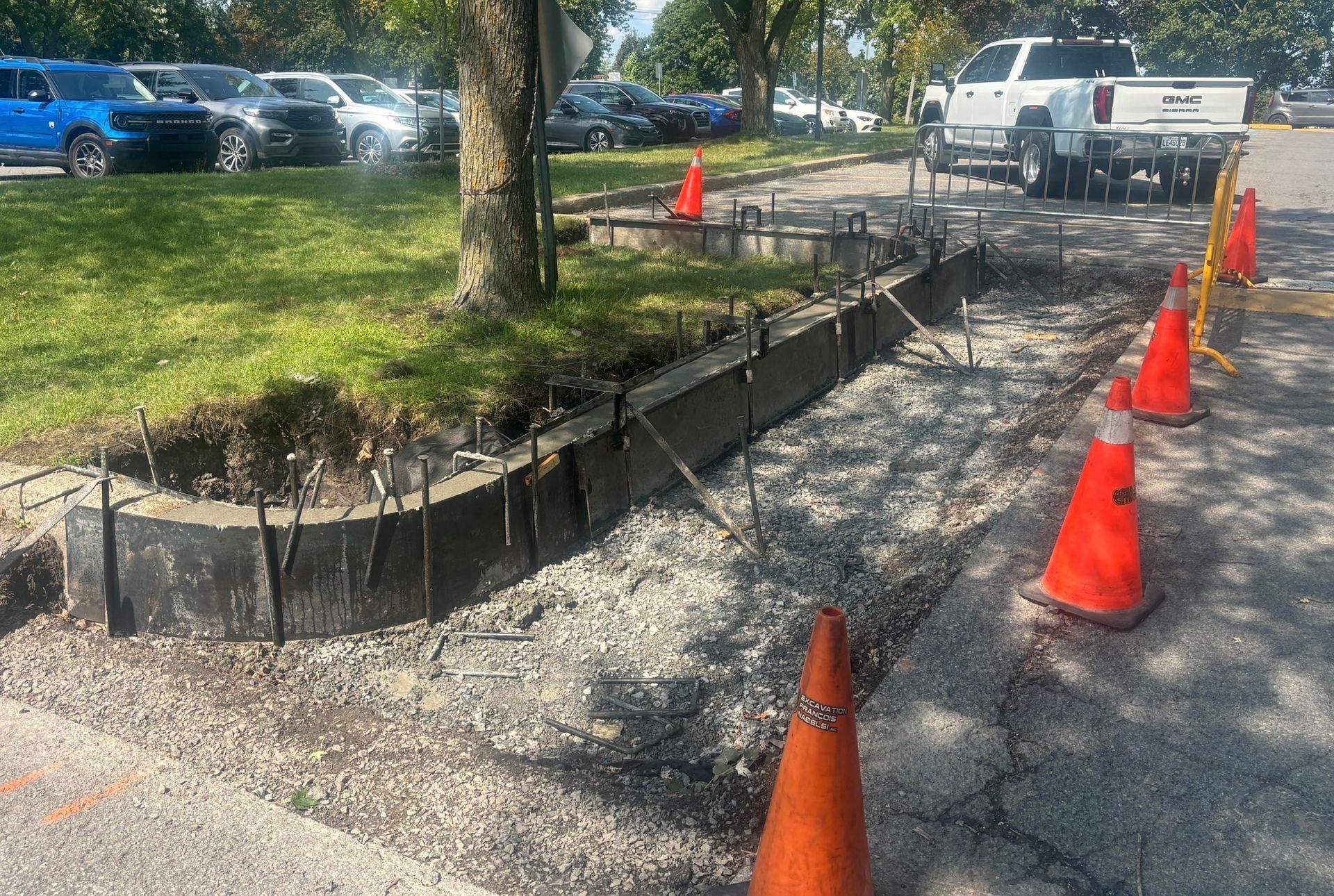 Bordure partiellement construite autour d'un arbre, avec des cônes orange signalant une zone de travaux en gravier près de voitures stationnées